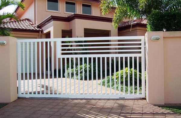 White aluminium slat sliding gate across a paved driveway, set between rendered pillars